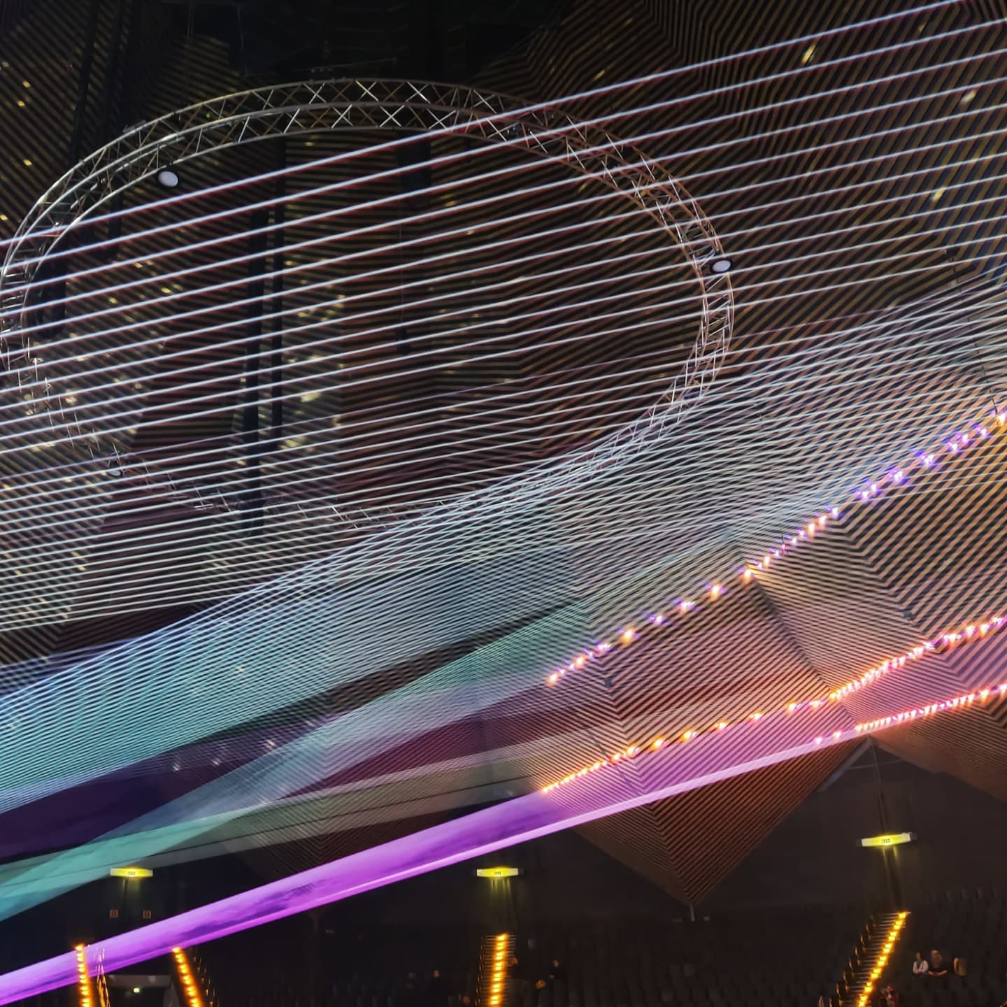 photo of a laser show during a concert. in the background, empty seats are visible, the laser show being some white horizontal lines, multiple big green to blue gradient and a purple stripe at the bottom